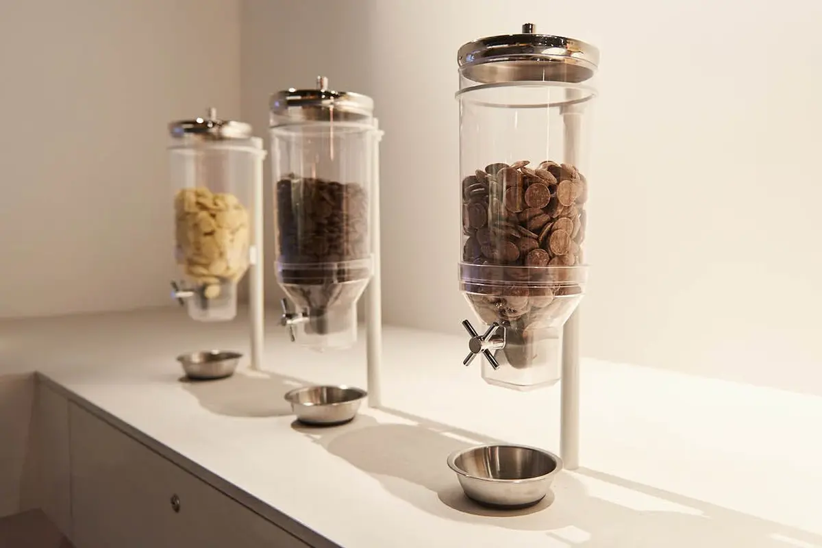 Trois distributeurs cylindriques contenant différentes céréales avec des bols en métal sous chacun d'eux, placés sur un comptoir blanc.