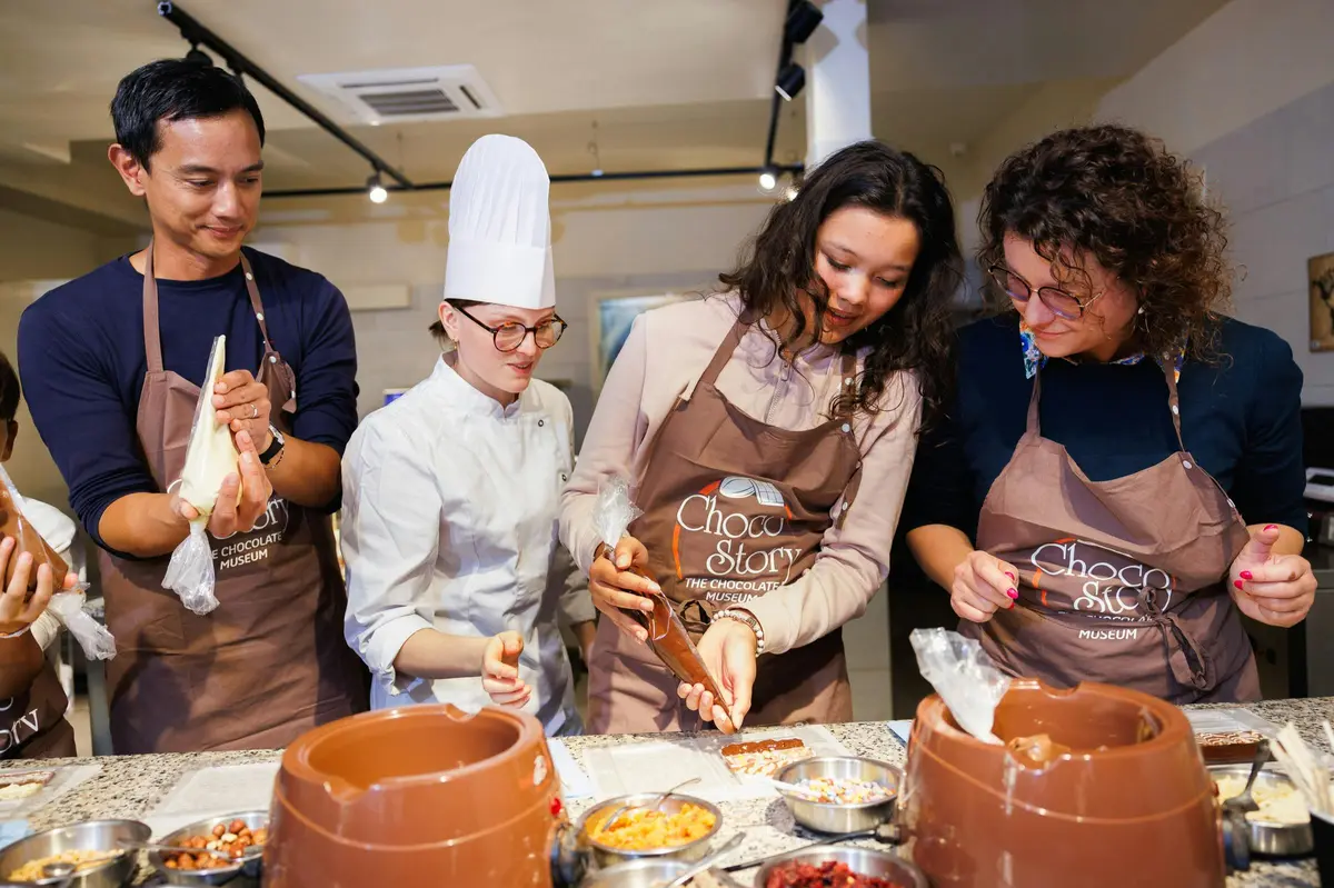 Un groupe de personnes en tablier et un chef décorent des chocolats dans un atelier. Des bols d'ingrédients sont posés sur la table.