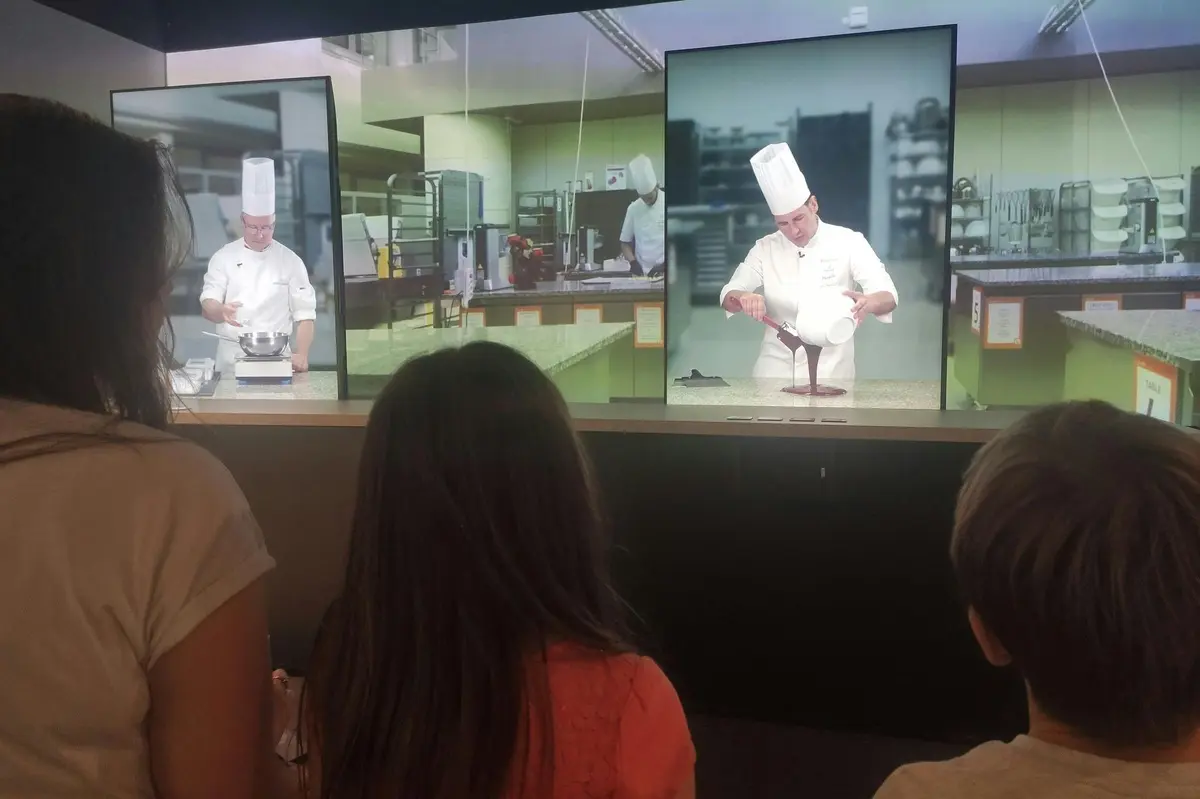 Deux personnes regardent des chefs sur des écrans dans une cuisine, faisant des démonstrations de techniques culinaires en uniforme blanc et chapeau haut de forme.