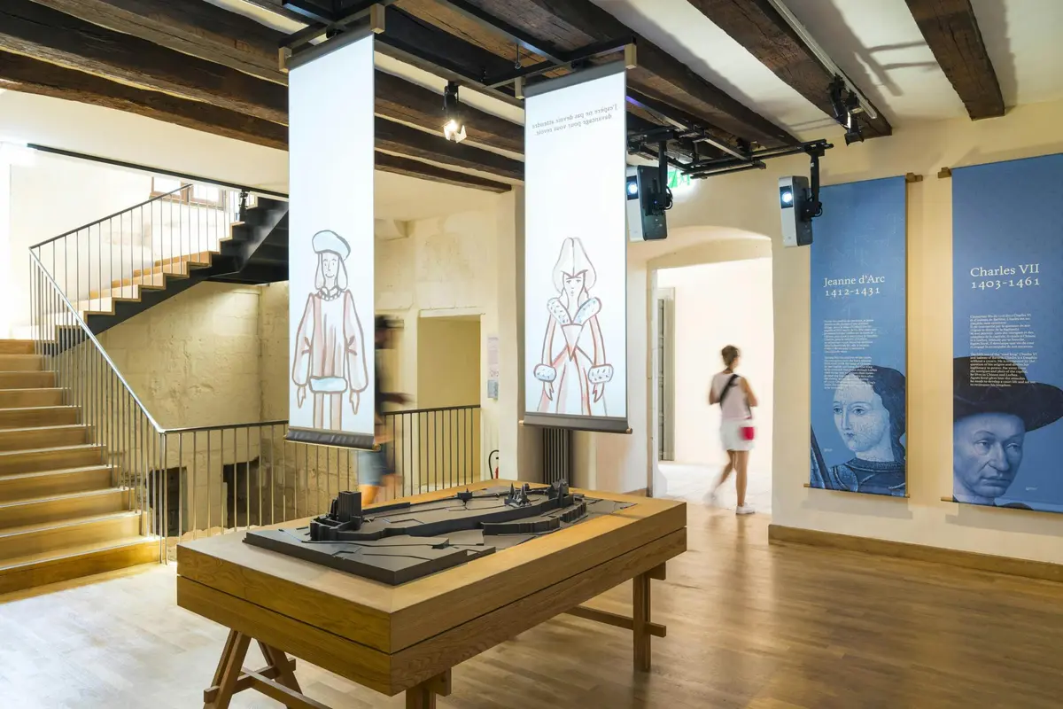 Exposition dans un musée présentant les bannières de Jeanne d'Arc et de Charles VII, avec une table de présentation en 3D et un visiteur qui passe devant.