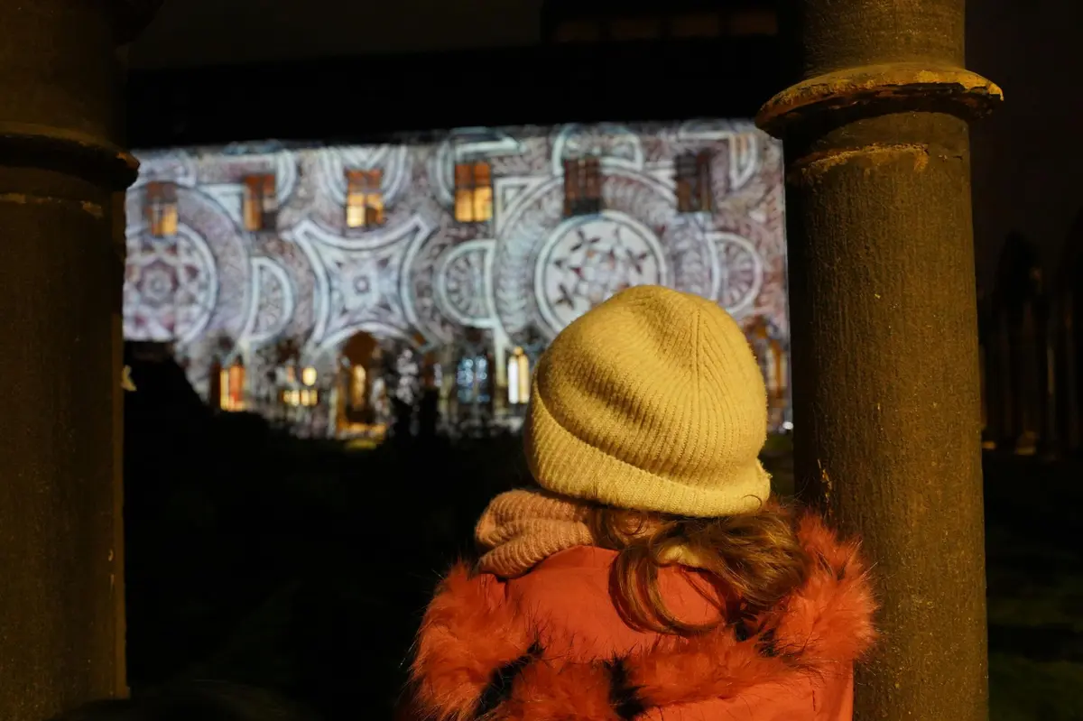Un enfant, coiffé d'un bonnet jaune et vêtu d'un manteau rouge, observe un immeuble illuminé la nuit par des projections lumineuses aux motifs variés.
