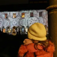 Un enfant, coiffé d'un bonnet jaune et vêtu d'un manteau rouge, observe un immeuble illuminé la nuit par des projections lumineuses aux motifs variés. &copy; Musée Unterlinden