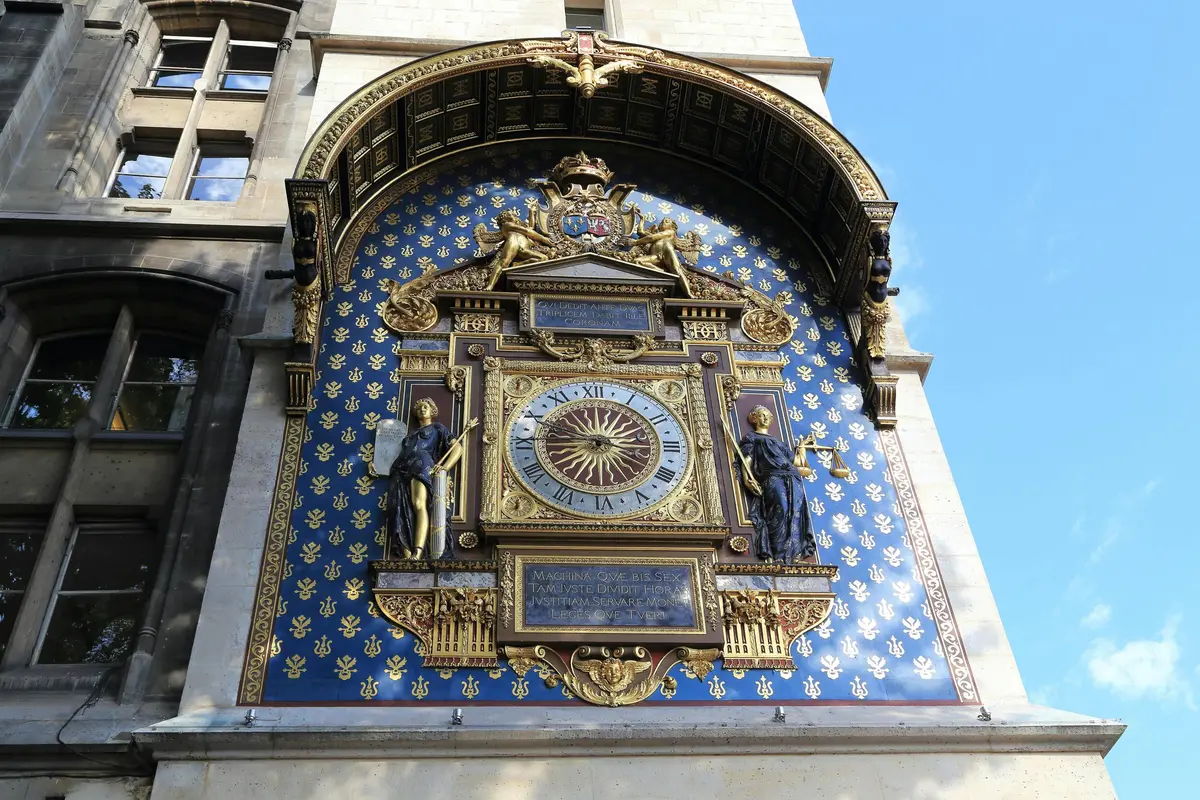 Horloge ornée sur une façade de bâtiment avec des détails complexes, deux statues, un fond bleu avec des motifs de fleurs de lys d'or.