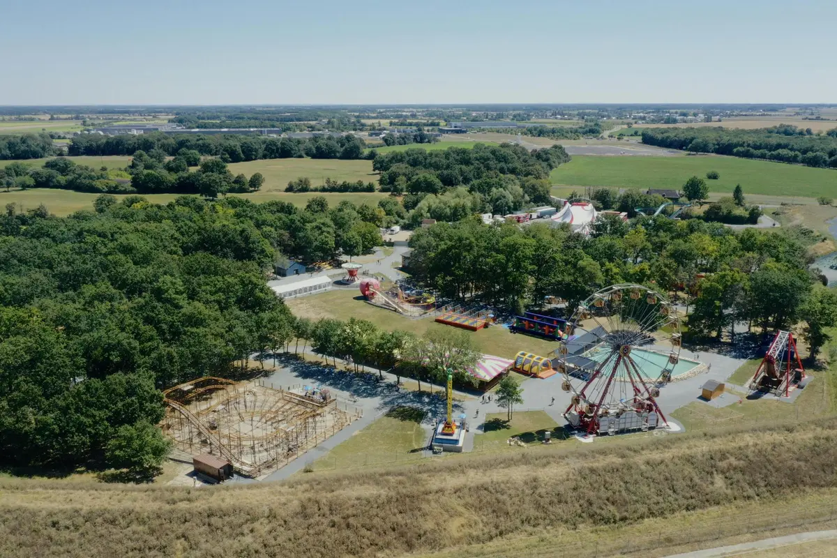 Vue aérienne d'un parc d'attractions de campagne avec une grande roue, des montagnes russes et d'autres manèges entourés d'arbres et de champs.