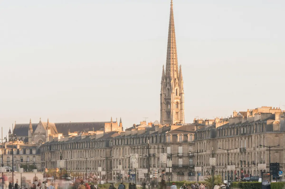 Un paysage urbain comprenant des bâtiments historiques et une haute flèche d'église sous un ciel clair, avec des personnes au premier plan.