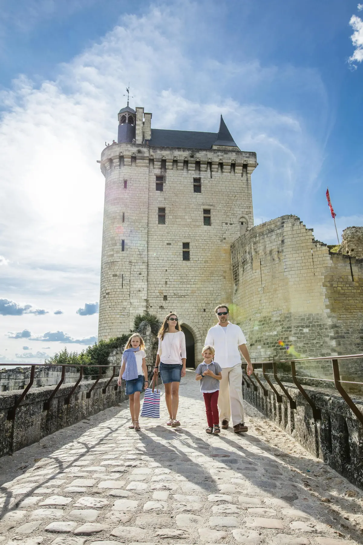 Une famille de quatre personnes traverse un pont de pierre en direction d'un grand château médiéval sous un ciel partiellement nuageux.
