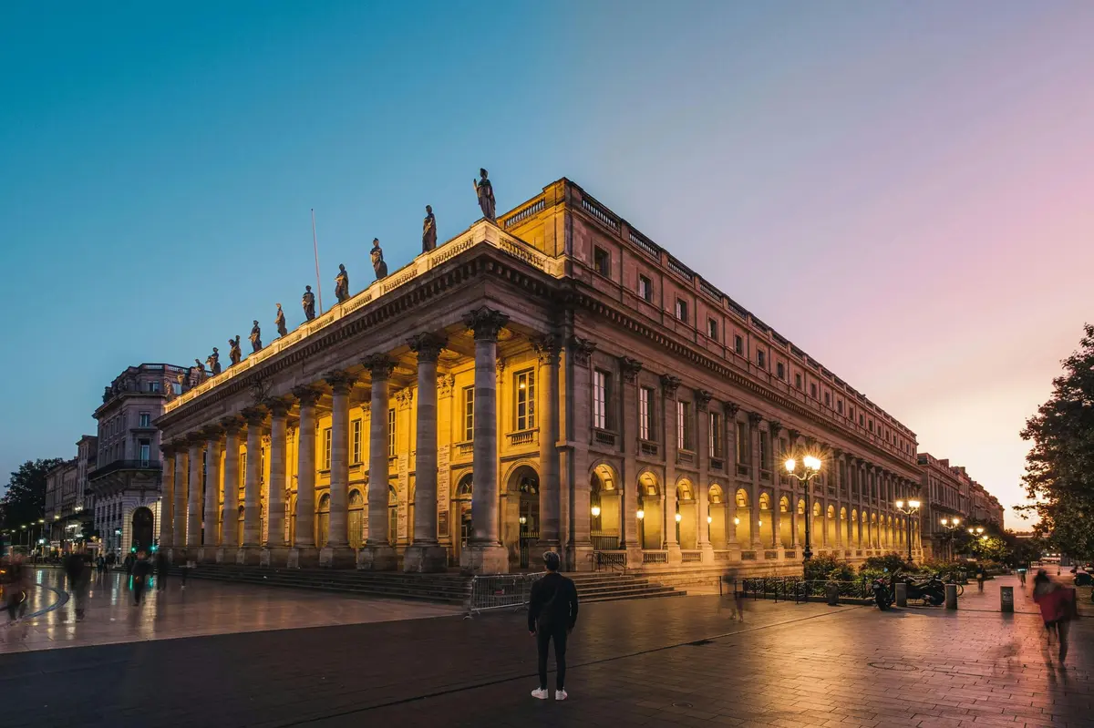 Une personne se tient devant un bâtiment néoclassique illuminé, orné de colonnes et de statues, à la tombée de la nuit.