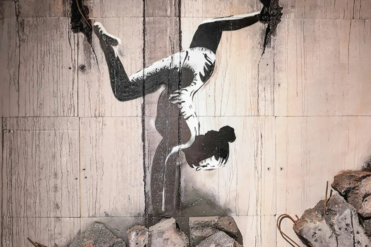 Une illustration réalisée au pochoir représentant une personne faisant le poirier, peinte sur un mur dont la partie inférieure est recouverte de gravats et de débris.