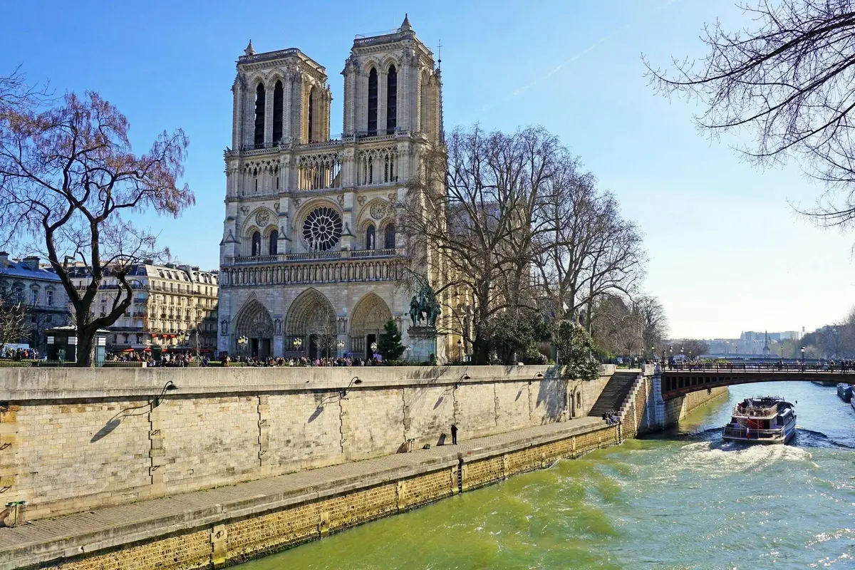 La cathédrale Notre-Dame avec ses tours jumelles, une rivière verte au premier plan, des arbres sans feuilles et un ciel bleu clair.