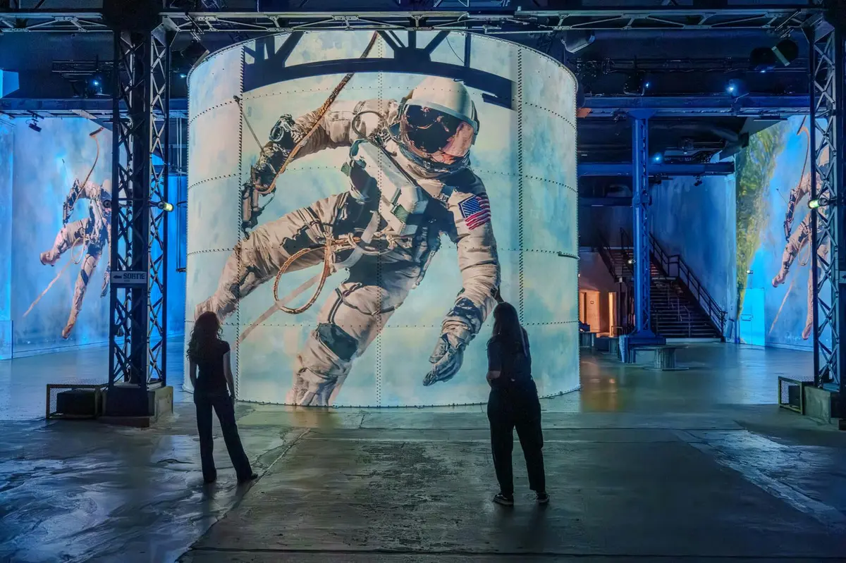 Deux personnes se tiennent devant un grand écran cylindrique représentant un astronaute flottant dans l'espace à l'intérieur d'une pièce faiblement éclairée.