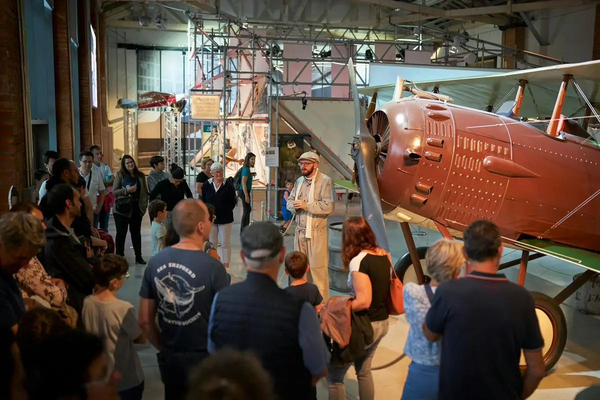 Un groupe de personnes écoutant un conférencier devant un avion d'époque dans un musée couvert.