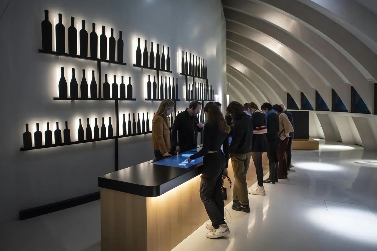 Des personnes examinent un écran tactile dans une salle de dégustation de vins moderne dont les murs sont ornés de silhouettes de bouteilles illuminées.