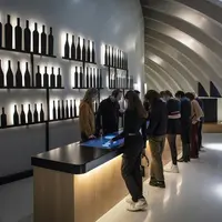 Des personnes examinent un écran tactile dans une salle de dégustation de vins moderne dont les murs sont ornés de silhouettes de bouteilles illuminées. &copy; ®Photo Anaka - Agence Clémence Farrell_Illusio_XTU architects