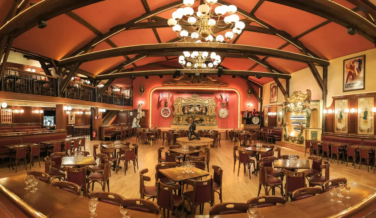 Grande salle à manger ornée de tables et de chaises en bois, de lustres, d'une scène au décor élaboré et d'une mezzanine.