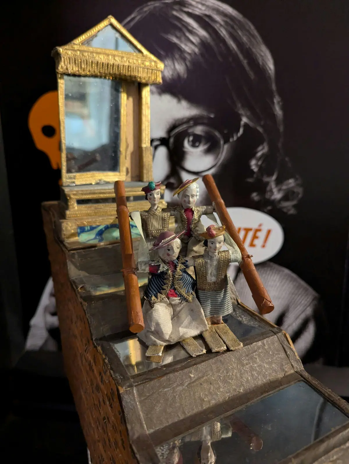 Quatre petites figurines en tenue décorative tenant des perches, avec une structure réfléchissante derrière elles. Un grand visage à l'arrière-plan.