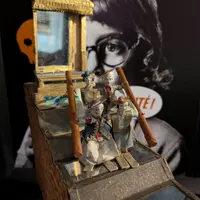 Quatre petites figurines en tenue décorative tenant des perches, avec une structure réfléchissante derrière elles. Un grand visage à l'arrière-plan. &copy; Villa de Colmar