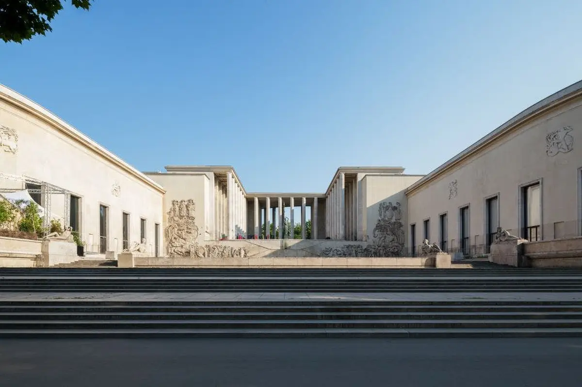 Un bâtiment néoclassique avec des colonnes, des sculptures décoratives et un large escalier sous un ciel bleu clair.