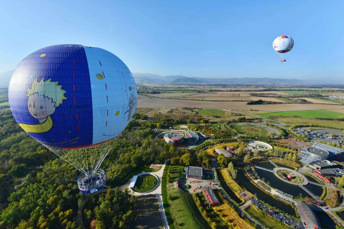 Deux montgolfières flottent au-dessus d'un parc avec des jardins, des cours d'eau et des bâtiments, entouré d'un vaste paysage.