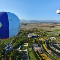 Deux montgolfières flottent au-dessus d'un parc avec des jardins, des cours d'eau et des bâtiments, entouré d'un vaste paysage. DR