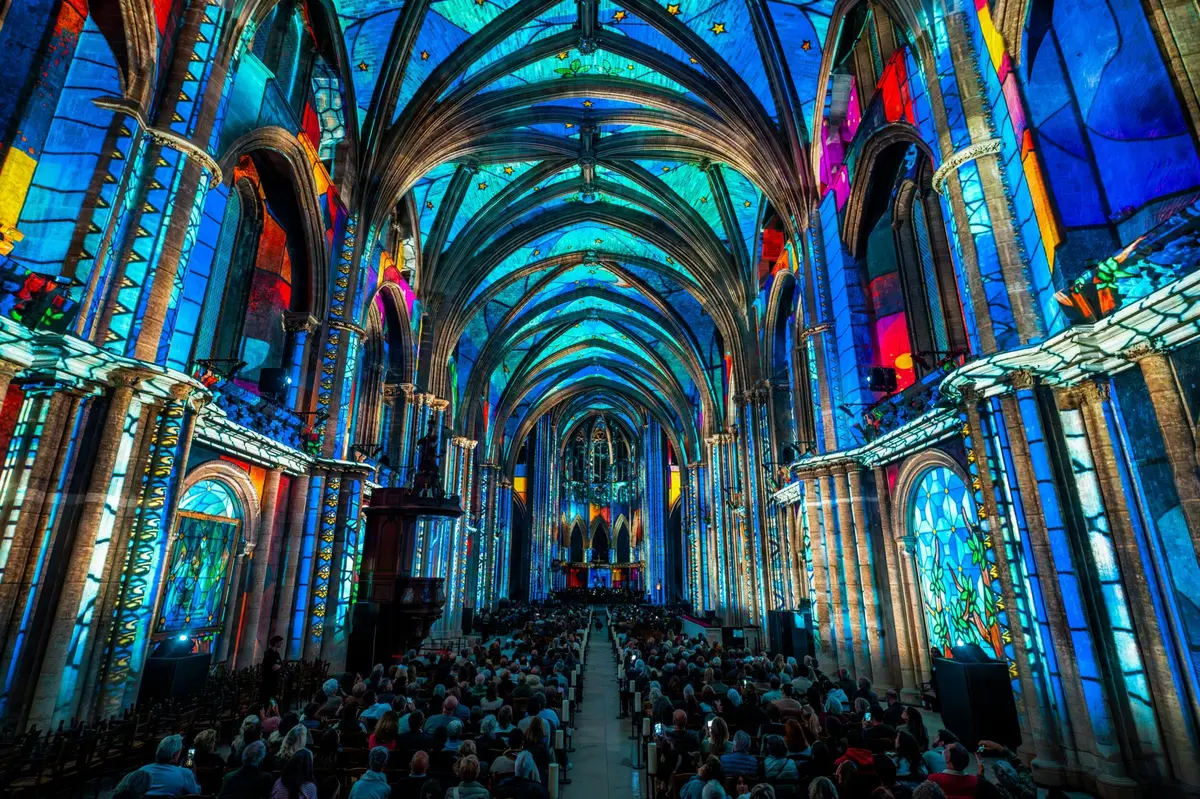 L'intérieur d'une cathédrale, avec ses vitraux colorés qui laissent passer la lumière, et une foule nombreuse assise le long de l'allée centrale.