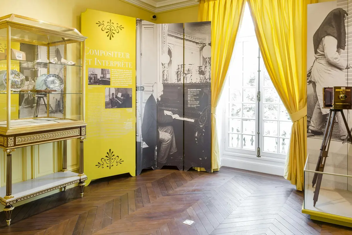 Une présentation de musée avec un panneau jaune, une cloison de photos historiques, une vitrine et de grands rideaux jaunes au-dessus d'une fenêtre.
