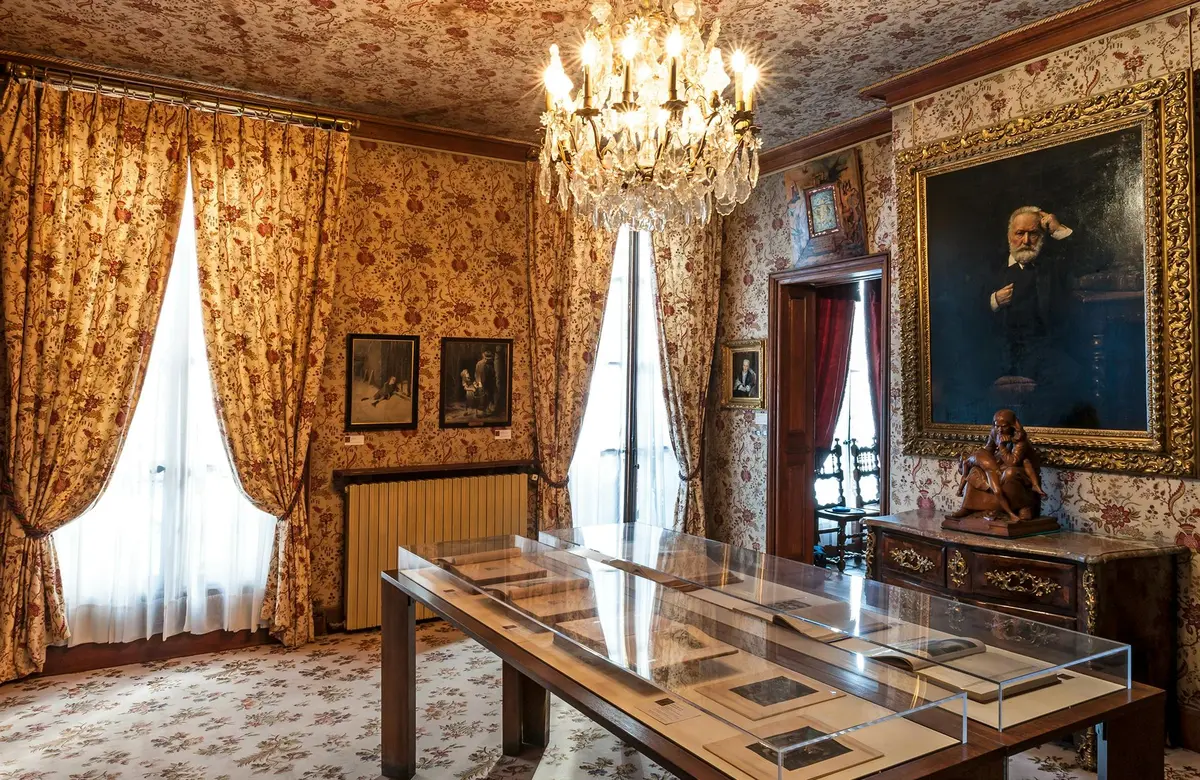 Pièce avec papier peint floral, lustre et œuvres d'art encadrées. Table avec vitrines au centre et meubles en bois ornés.
