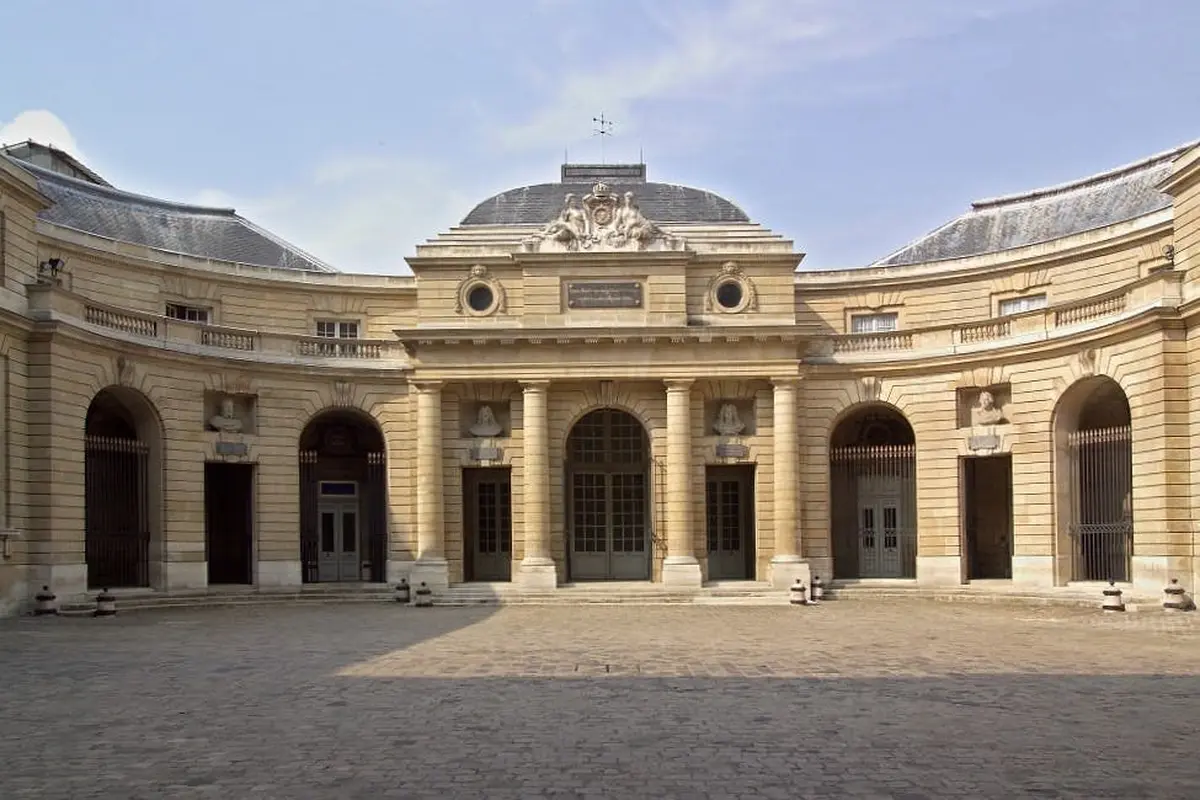Un bâtiment historique avec une façade incurvée, des fenêtres cintrées et des colonnes, situé dans une cour pavée sous un ciel clair.