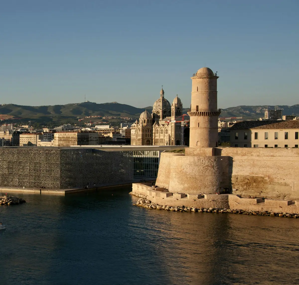 Un phare historique et un fort au bord de l'eau avec un bâtiment moderne et des montagnes en arrière-plan sous un ciel dégagé.