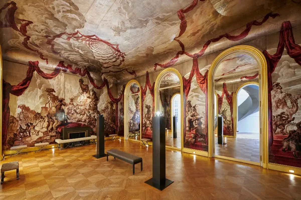 Salle ornée d'un grand plafond et de peintures murales, de miroirs encadrés d'or, d'un banc et de colonnes. Parquet et décor historique.