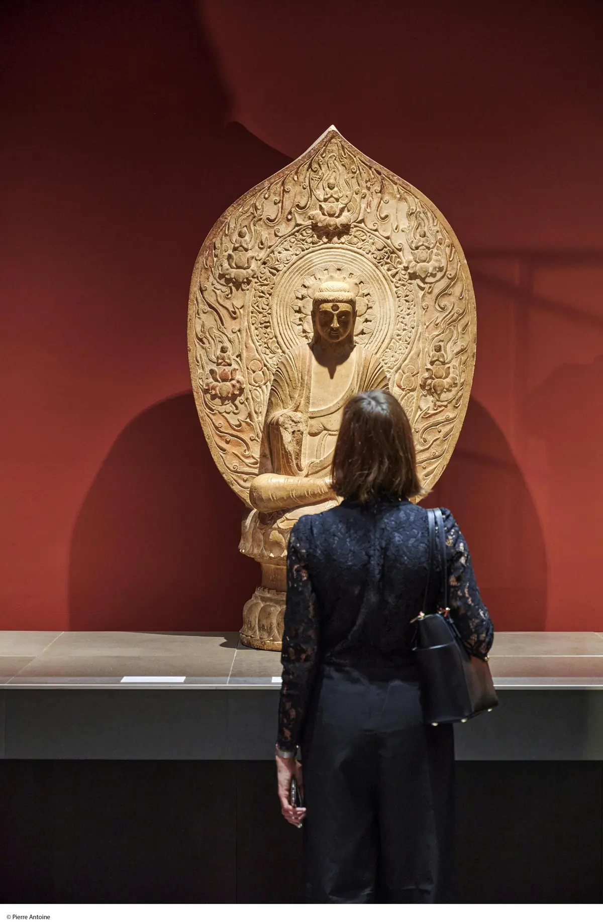 Une personne se tient devant une statue de Bouddha détaillée exposée dans un musée sur fond rouge.