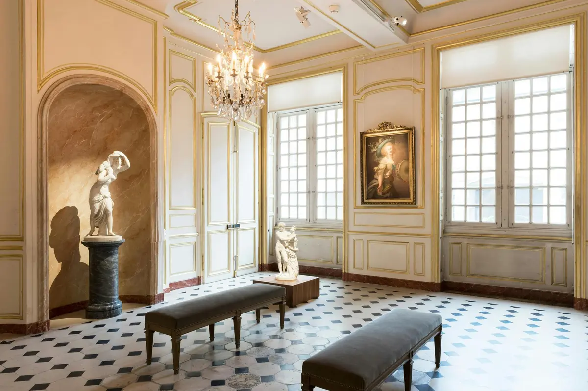 Salle élégante avec des lustres, des statues, un portrait encadré, des bancs et des carreaux de marbre à carreaux. De grandes fenêtres laissent entrer la lumière naturelle.