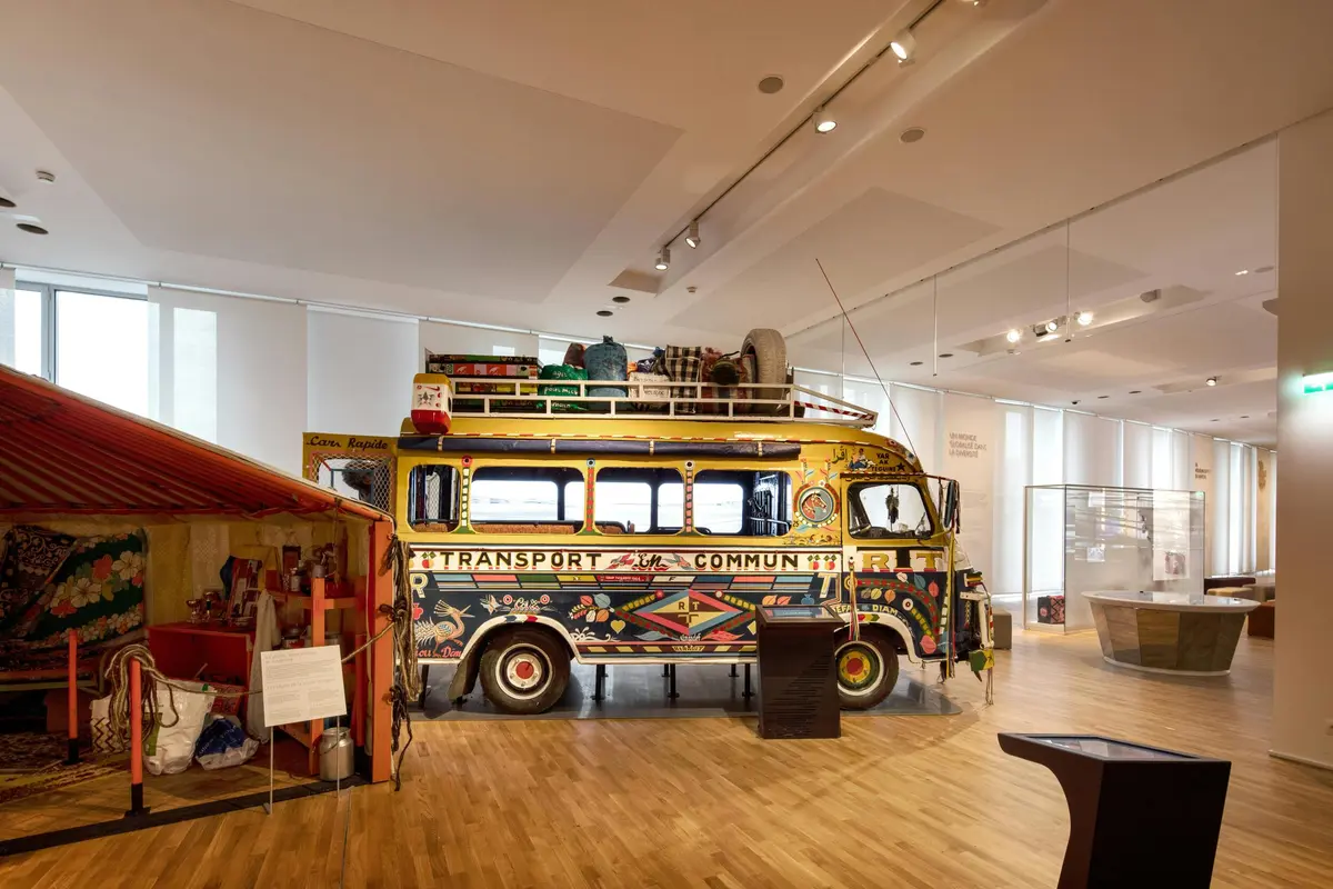 Un bus ancien décoré avec des bagages sur le toit, exposé dans une salle de musée lumineuse avec des planchers en bois.