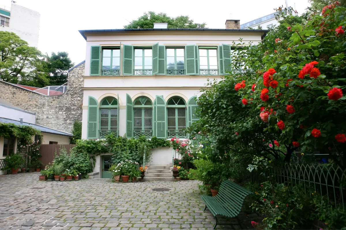 Une maison à deux étages avec des volets verts et des fenêtres cintrées, entourée d'un jardin avec des fleurs et un banc vert.