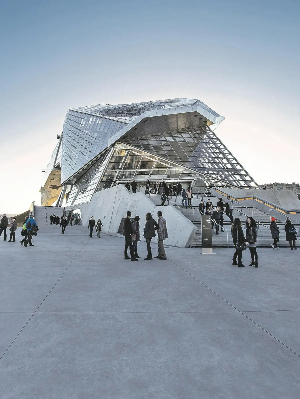 Les gens se promènent et se rassemblent autour d'un bâtiment moderne et anguleux en verre avec de grands escaliers en béton sous un ciel clair.