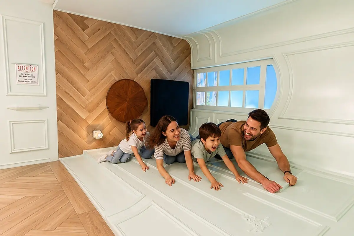 Une famille de quatre personnes s'amuse dans une pièce inclinée aux murs recouverts de panneaux blancs et dotée d'une fenêtre, ce qui donne l'illusion de grimper.