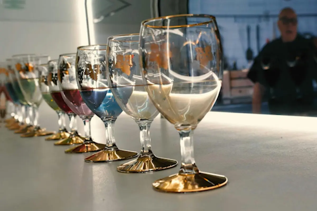 Une rangée de six verres remplis de liquides de différentes couleurs, chacun avec des éléments en or, disposés sur une surface réfléchissante.