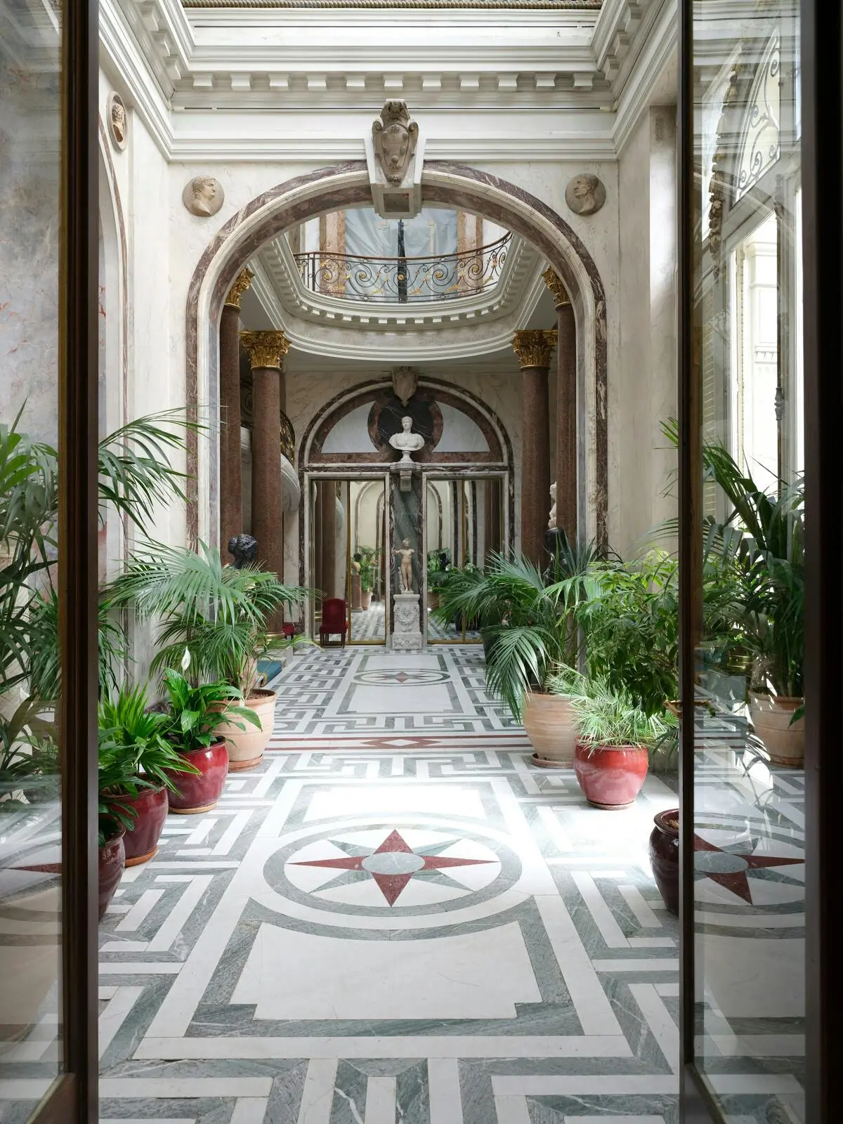 Couloir orné de colonnes de marbre, d'une statue au centre et de plantes en pot le long du sol à motifs.