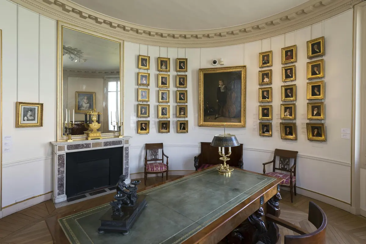 Pièce élégante avec une table en cuir vert, un grand miroir au-dessus de la cheminée et de nombreux portraits dans des cadres dorés sur les murs.