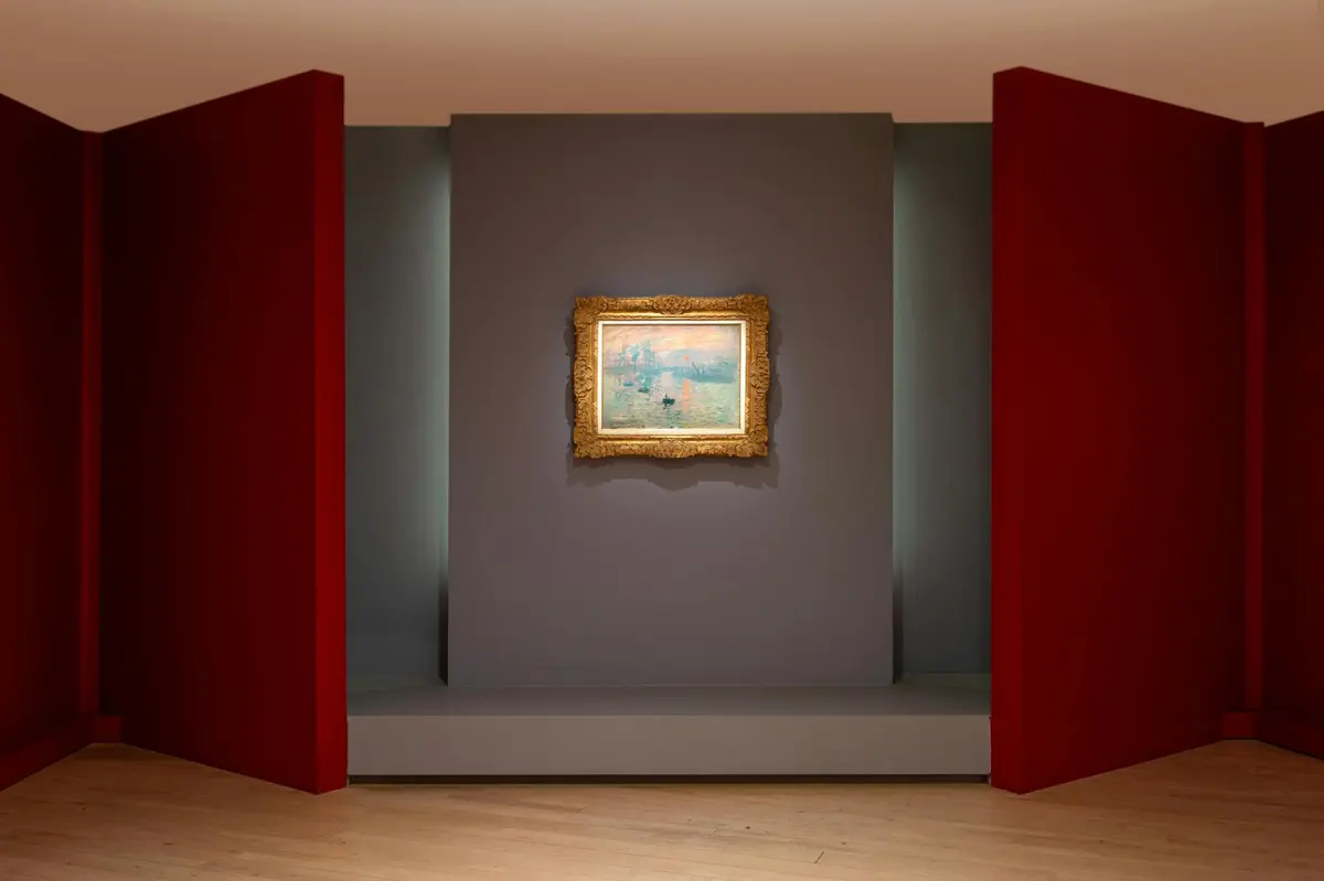 Un tableau encadré accroché à un mur gris, encadré de deux panneaux rouges, dans une galerie dotée d'un parquet.