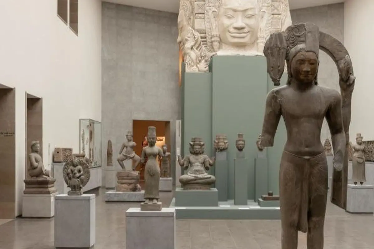 Une exposition muséale présentant des sculptures anciennes, dont une grande tête sculptée et plusieurs statues plus petites sur des piédestaux.