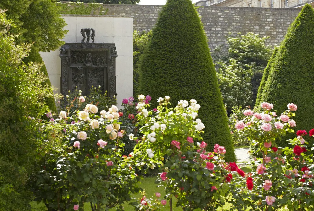 Jardin avec des roses roses, rouges et blanches en fleurs, des arbres coniques bien entretenus et un mur de pierre au relief sombre et orné.