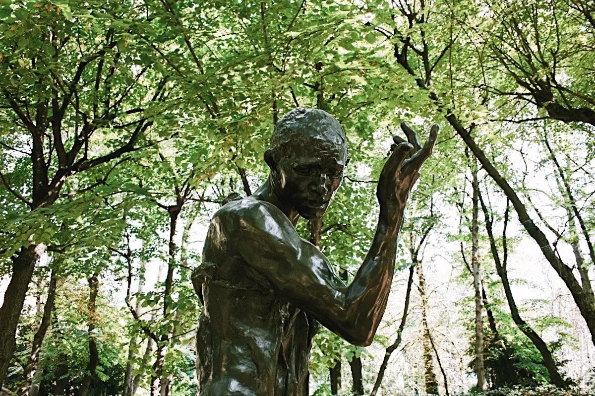 Statue en bronze représentant un personnage debout, les bras levés, sur fond d'arbres verdoyants.