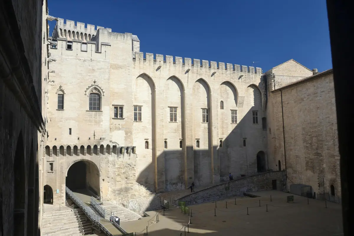 Cour historique en pierre avec une arcade et des rangées de fenêtres cintrées sur un bâtiment ressemblant à un château sous un ciel bleu clair.