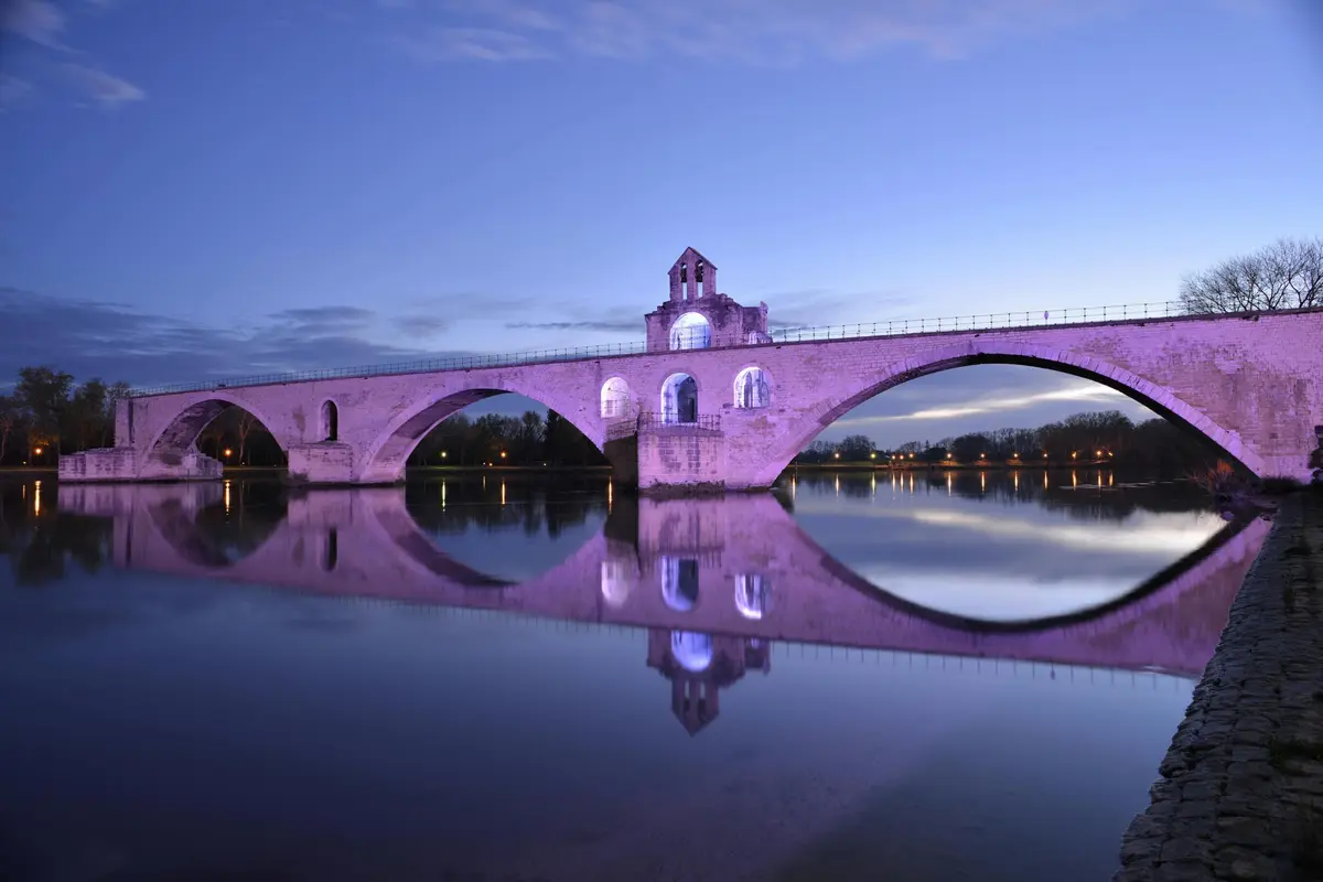 Pont en pierre avec des arches et une tour, illuminé en violet, se reflétant dans une rivière calme au crépuscule.