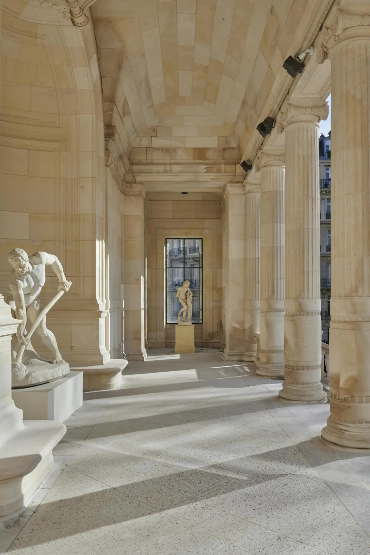 Une colonnade ensoleillée avec des statues classiques, dont un homme penché avec un outil et un personnage debout éloigné.