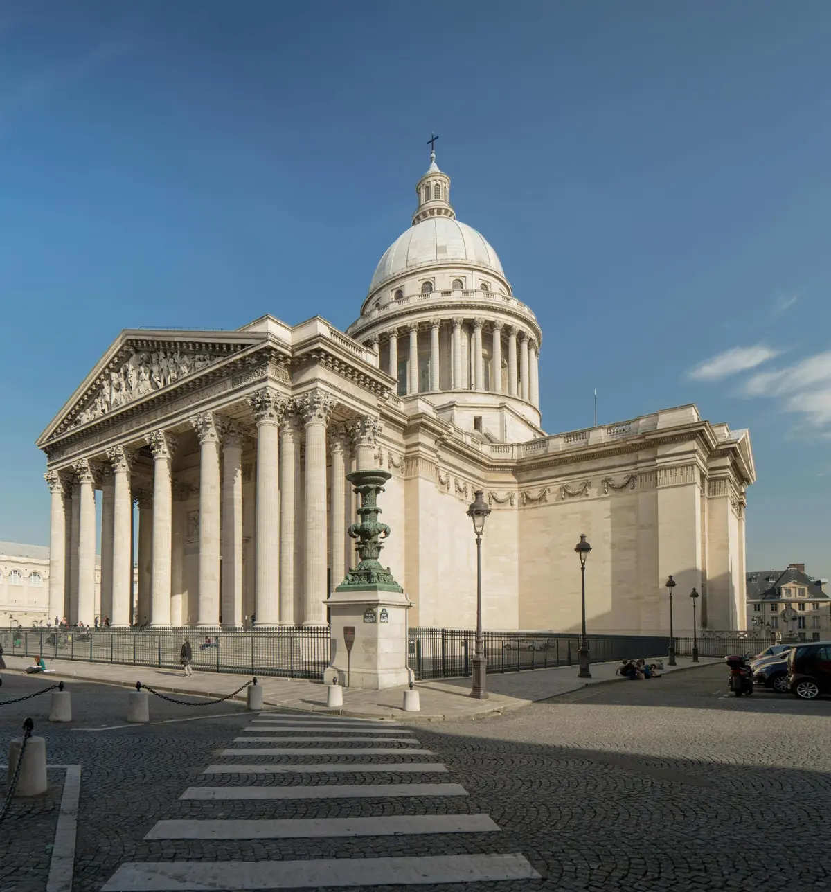 Un bâtiment néoclassique avec de grandes colonnes et un dôme sous un ciel bleu clair, vu d'un passage piéton dans une rue pavée.