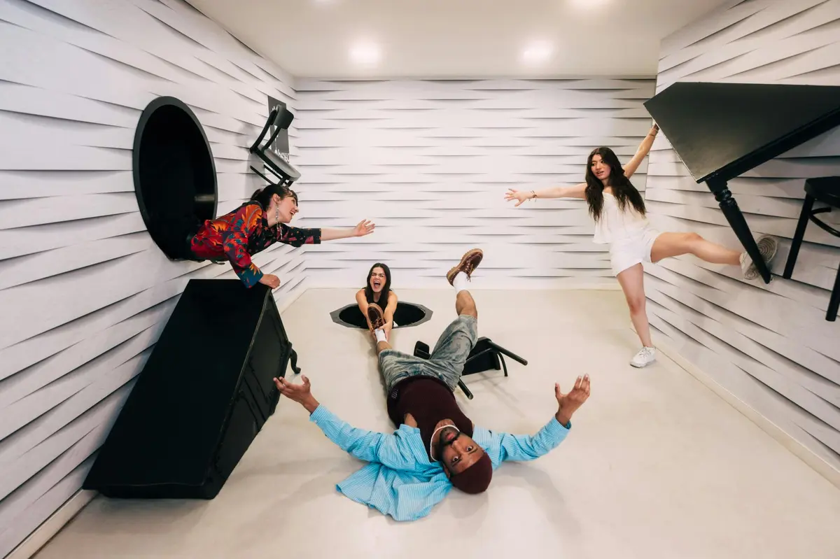 Quatre personnes interagissent dans une pièce ludique et inclinée avec des illusions d'optique, avec un mur conçu pour ressembler à un sol.