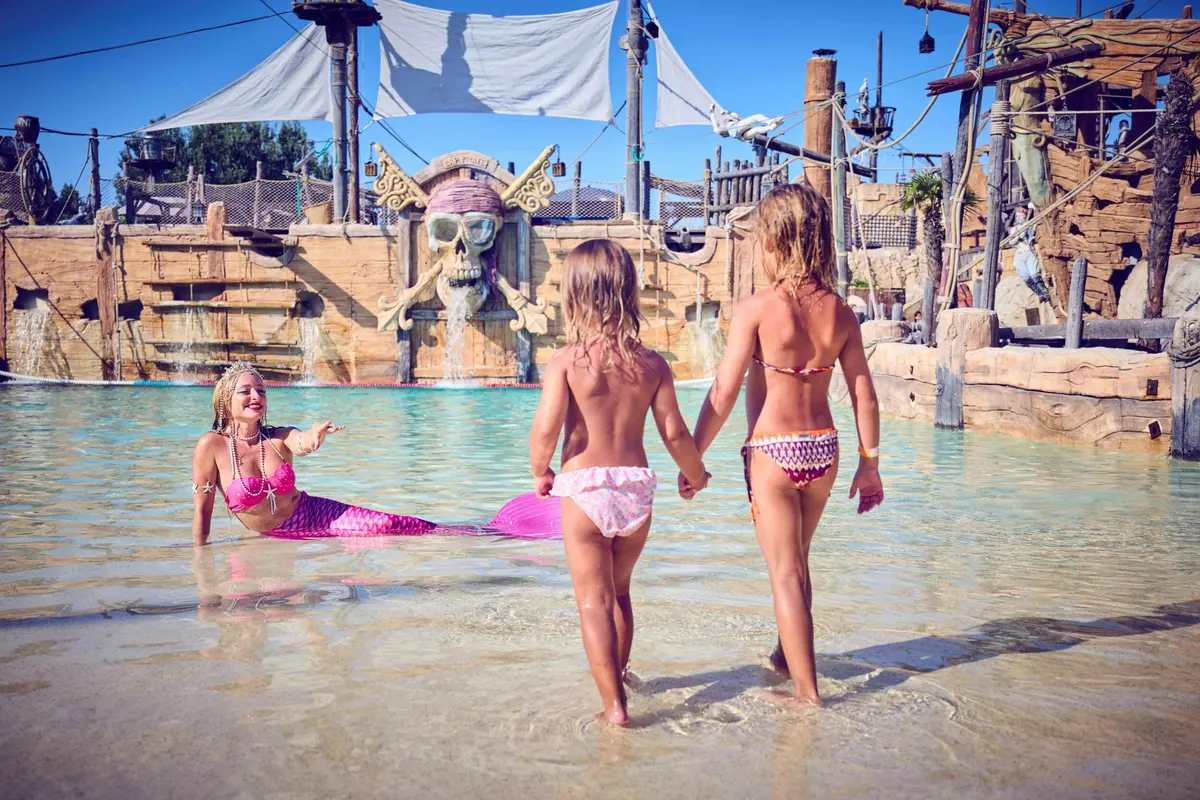 Deux enfants se tiennent dans l'eau peu profonde face à un artiste déguisé en sirène, près d'une toile de fond sur le thème des pirates.