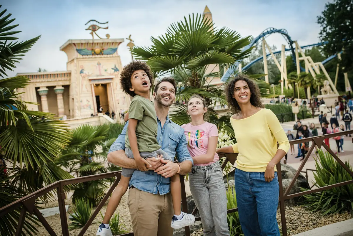 A smiling family of four in casual attire stands outdoors near palm trees, with an amusement park ride and themed building in the background.
