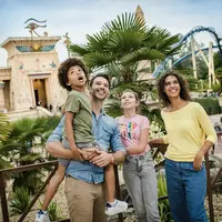 A smiling family of four in casual attire stands outdoors near palm trees, with an amusement park ride and themed building in the background. &copy; @sylvaincambon